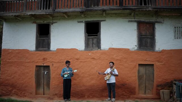 Two musicians perform outdoors in front of a traditional orange and white Himalayan mud house holding a ukulele and a carved sarangi