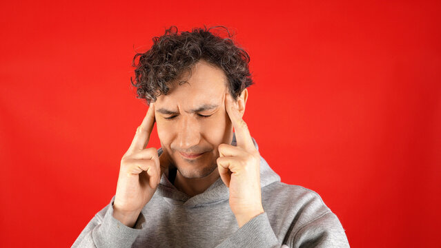 Young man grey hoodie pressing fingers temples grimacing severe headache pain migraine stress tension anxiety mental exhaustion isolated red studio background