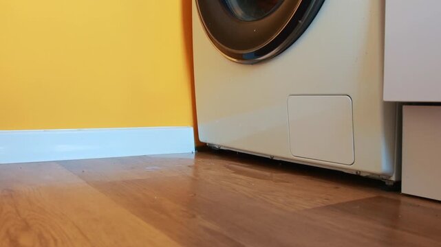 Water leaking from under a white washing machine onto a wooden laminate floor in a domestic room. Water leakage on the floor near a washing machine in the bathroom