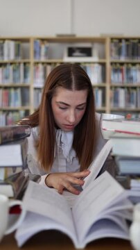 Vertical screen focused female student reads textbooks and drinks coffee while studying intensely in a library surrounded by books during demanding exam preparation alone