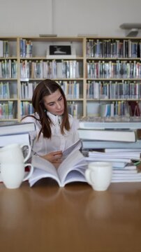 Vertical screen bored young woman slowly turns pages of a book at library table surrounded by large book stacks while losing interest and struggling to continue reading now