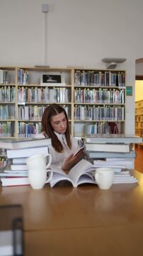 Vertical screen serious young woman reads a book at library table surrounded by stacks of books while focusing on information and continuing an intensive study period now