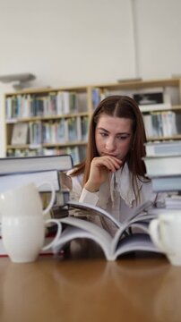 Vertical screen thoughtful young woman reads an open book in library resting hand near her mouth while concentrating on difficult study material and searching for answers