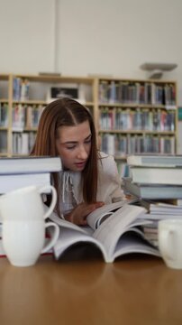 Vertical screen focused young woman leans over open books in a library reading carefully and searching for useful information while surrounded by stacks of study material