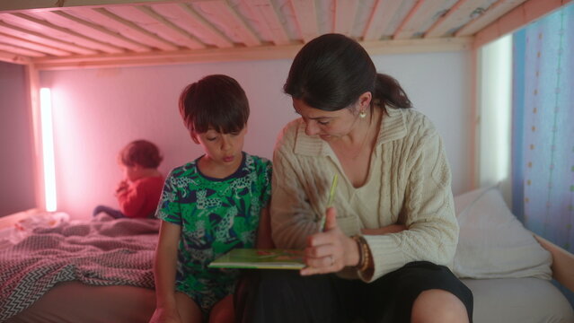 Mother helping son sound out words from storybook before bedtime while toddler relaxes in background creating nurturing family atmosphere focused on literacy and childhood learning
