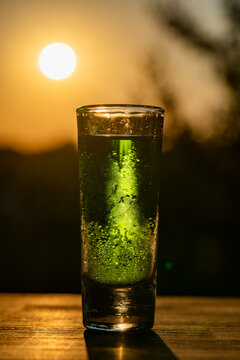 Green alcohol shot glass at sunset background. Tall shot glass filled with bright green alcoholic drink or liqueur covered in condensation drops standing on wooden table against golden sunset sky