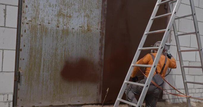 Industrial painter wearing work overalls and a cap using a spray gun to apply brown primer on a rusty metal gate, performing corrosion protection and surface renovation work on a construction site.