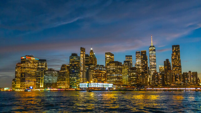 New York City skyline glowing at dusk, with illuminated skyscrapers, waterfront reflections, and a dramatic evening sky creating a vibrant urban scene for travel and city lifestyle content.