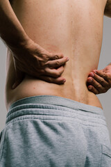 Naklejka premium A man with a summer tan massages his sore lower back with his fingers. A guy in gray shorts, turning away from the camera, scratches his pimply back. men's health: prevention of spinal diseases