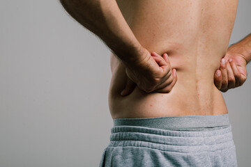 Naklejka premium A man with a summer tan massages his sore lower back with his fingers. A guy in gray shorts, turning away from the camera, scratches his pimply back. men's health: prevention of spinal diseases
