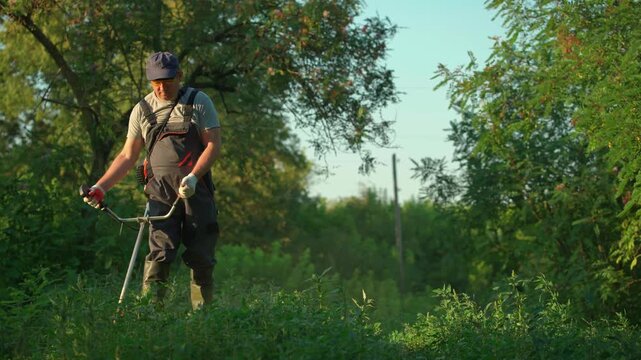 Gardener in gray shirt and overalls operates a string trimmer in a lush green field, surrounded by trees and foliage, demonstrating the process of clearing grass and weeds in a natural outdoor setting
