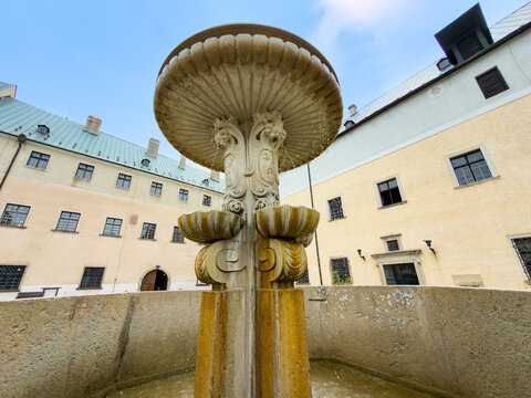 Carved stone fountain column at Cerveny Kamen Castle courtyard. Central column with ornate water spouts and bas reliefs in historic courtyard under sunlight.