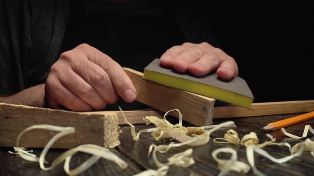 Skilled craftsman uses sanding block on wooden piece, focusing on smoothing edges while surrounded by wood shavings, pencil, and additional wood pieces on a dark work surface