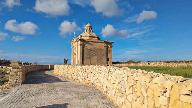 Kappella San Nikolaw chapel beside paved path and traditional Maltese stone walls, green fields and bright blue sky with scattered clouds in rural Malta