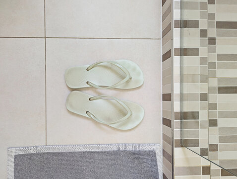 Mint flip flops on neutral tiled bathroom floor beside glass shower enclosure and gray bath mat, clean simple home interior