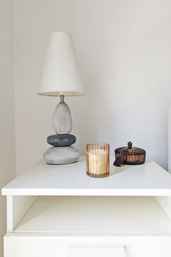Stone table lamp, glowing ribbed candle holder and dark glass decorative container on white cabinet against neutral wall