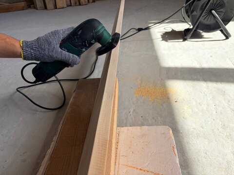 Worker sanding the edge of a wooden plank with an electric detail sander. A construction or renovation site