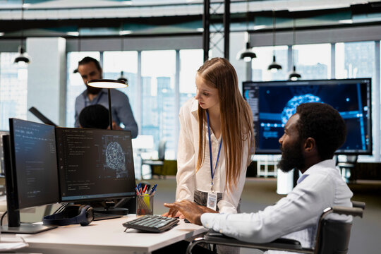Upbeat technician with a disability and a young junior developer use AI scripts to automate tasks in an inclusive office, including feature implementation by a worker in a wheelchair.