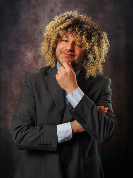 Man with curly hair, suit, thoughtful pose. Hipster male in his 40s or 50s. Dark background, studio lighting. Smiling, hand near chin. Professional portrait style.