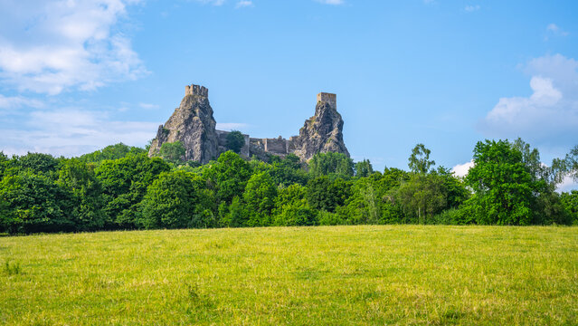 Trosky castle ruins rise above a green field in Bohemian Paradise. The structure is surrounded by trees and set against a blue sky. The scene shows nature and history together.