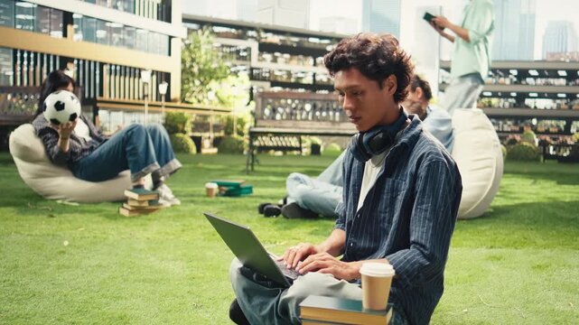Male student smiles and writes notes on a laptop from the grass at a college campus, with a coffee cup and stacked books nearby for independent study, exam preparation and outdoor learning.