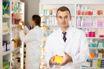 Fototapeta premium Man pharmacy employee presents box, offers to share information about remedy for problem skin. Male pharmacist will prompt and answer question, help determine choice