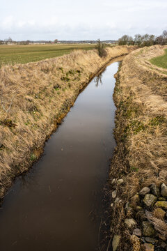 Narrow rural drainage canal winding through dry grass farmland under overcast sky.