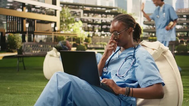 The portrait shows a woman student smiling and typing on a laptop from a beanbag in a modern outdoor campus courtyard, with a med school peer nearby in blue scrubs and a stethoscope.