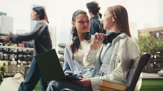 Women classmates collaborate on a laptop while seated on a bench at a university campus, discussing coursework and taking notes for exam preparation, supporting peer tutoring and group study.
