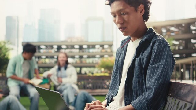 Focused international student checks notes on a laptop from a campus bench, with stacked books and a coffee cup nearby in a quiet independent outdoor learning session for exam preparation.
