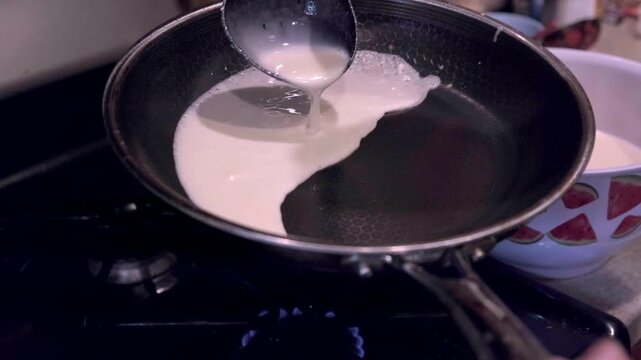 Pouring thin white batter from a ladle into a hot non-stick frying pan on a gas stove to make crepes