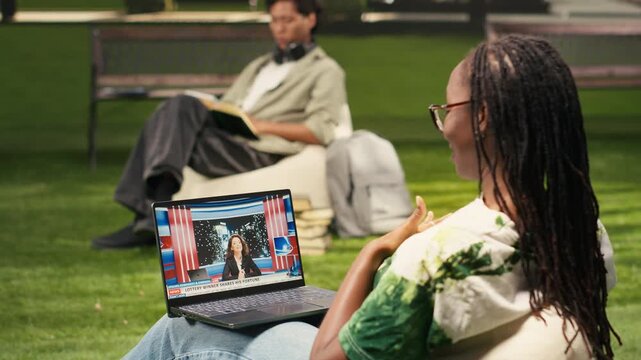The woman watches a news report segment about artificial intelligence on a laptop during outdoor campus study break, for digital media communication, for current affairs content, on a college campus.