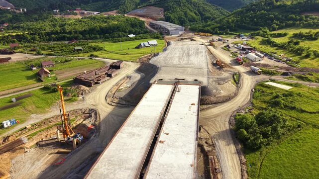 Aerial views of a major highway viaduct under construction across a broad mountain valley, showing completed concrete bridge deck sections, active earthworks, cranes and a nearby road interchange besi