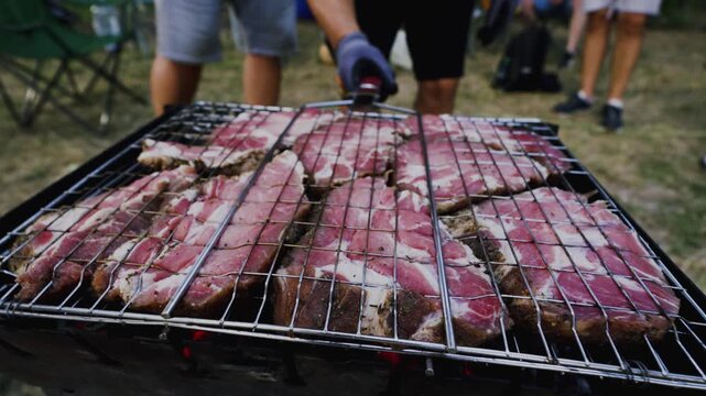 Grilling meat on a barbecue with visible flames and smoke, surrounded by people enjoying outdoor cooking in a grassy area during a summer gathering