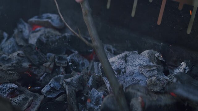 Charcoal grill with glowing embers and smoke as a person stirs the ashes with a stick, showcasing the progression of a campfire preparation in an outdoor setting