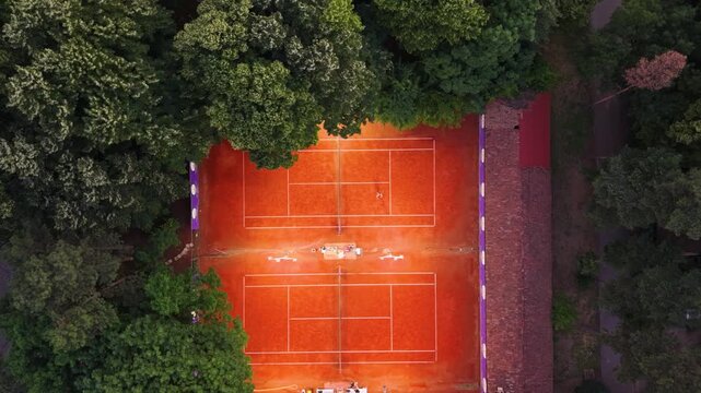 Top-down aerial view of a red clay tennis court surrounded by dense green trees, with crisp white court markings and warm sunlight creating a strong geometric sports pattern.