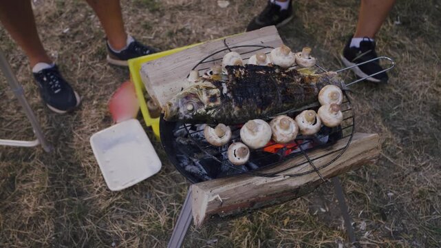 Grilled fish and mushrooms cooking on a charcoal grill outdoors, surrounded by grass and feet of people wearing casual footwear in a summer setting
