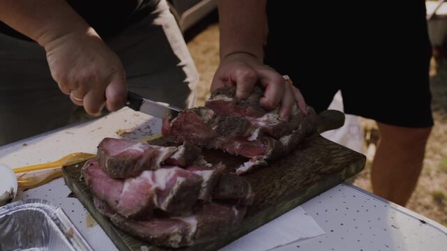 Close-up of hands slicing cooked meat on a wooden cutting board outdoors, showcasing the progression of cutting technique and meat texture in a casual setting