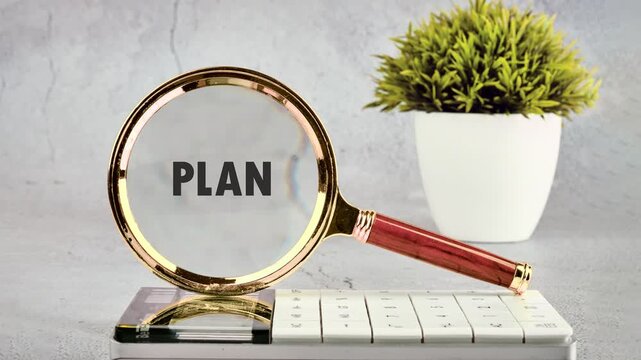 A person uses a magnifying glass to examine the word plan on a desk with a plant and keyboard. This happens in an office space