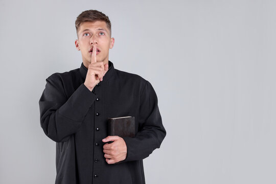 Priest in cassock with Holy Bible showing hush gesture on grey background. Space for text