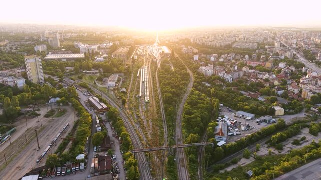 Aerial sunset view of a large railway yard and train depot in Sofia, Bulgaria, with numerous passenger trains, parallel tracks, maintenance buildings and rail infrastructure extending through the city