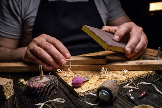 A craftsman is shaping wood pieces in a workshop. They hold a sanding block and are surrounded by various tools. Light highlights the workspace and the wood shavings