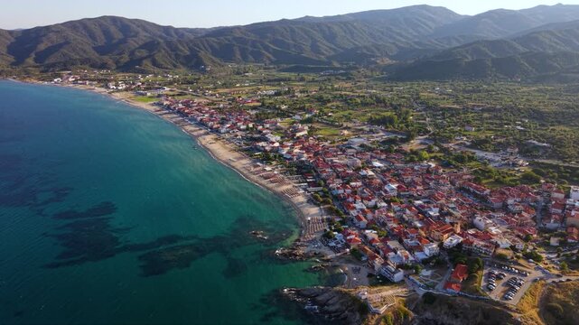 Aerial view of Toroni in Sithonia, Halkidiki, Greece, showing the coastal village beside a long sandy beach, clear turquoise Aegean Sea and rugged green hills in warm summer evening light.