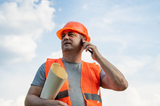 A worker in safety gear makes a phone call while holding construction plans. The setting is an outdoor construction site under a clear sky