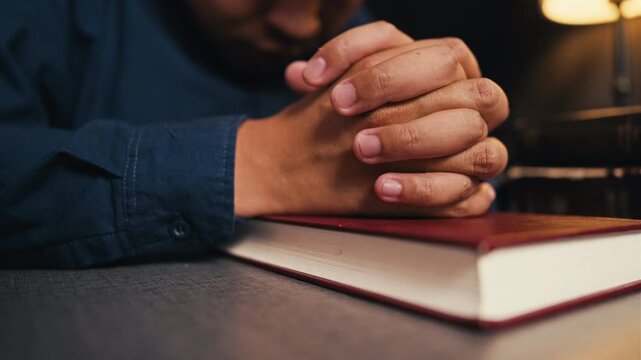 Close up of a religious man folded hands praying on the holy bible in quiet dark room, worshiping God with faith, devotion, spirituality, hope and belief for christian or catholic religion.
