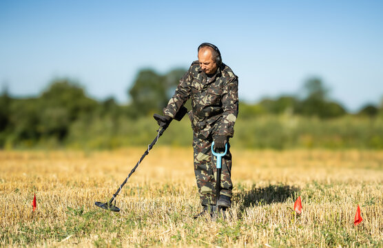 A treasure hunter dressed in camouflage stands in a field using a metal detector to find items underground. Nearby, small red flags mark points of interest. The sun is shining