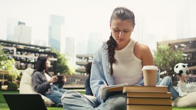 Focused student writes in a notebook beside an open book on a campus lawn, taking coursework notes during independent outdoor study.