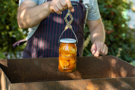A cook in a striped apron holds a jar full of canned tomatoes over a metal box. The scene takes place outside in a garden. Canning vegetables