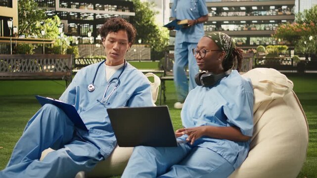 Diverse med school peers in blue scrubs collaborate over laptop notes while seated on beanbags, studying coursework in a modern outdoor campus setting.