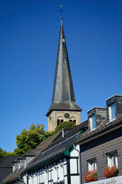 Tall slate church spire of St Walburga towering over timber-framed and slate-clad houses in Overath. Clear blue sky.